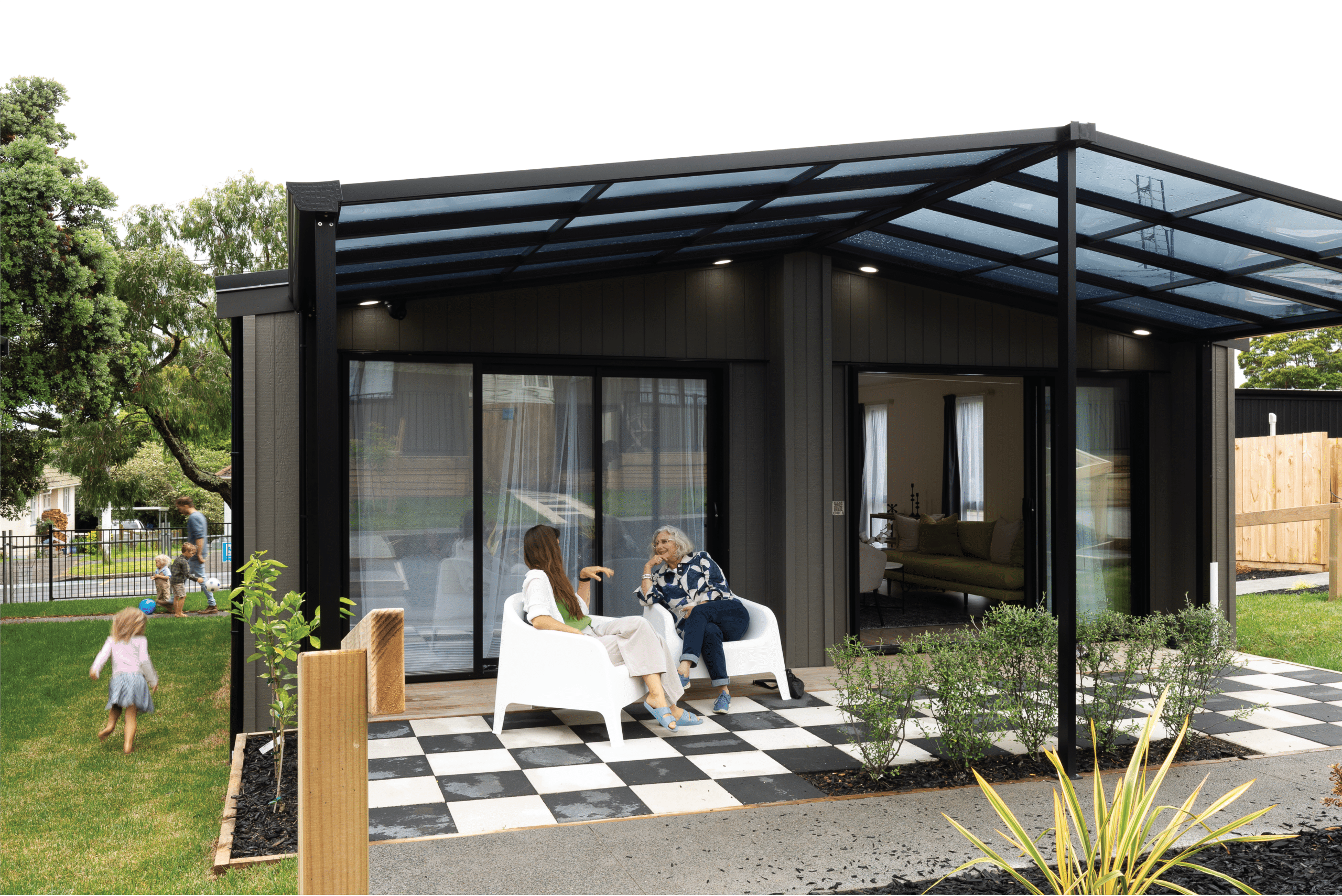 Photo with a mum and grandma speaking under a pergola outside an ecopod home, while a child runs towards her dad outside.  