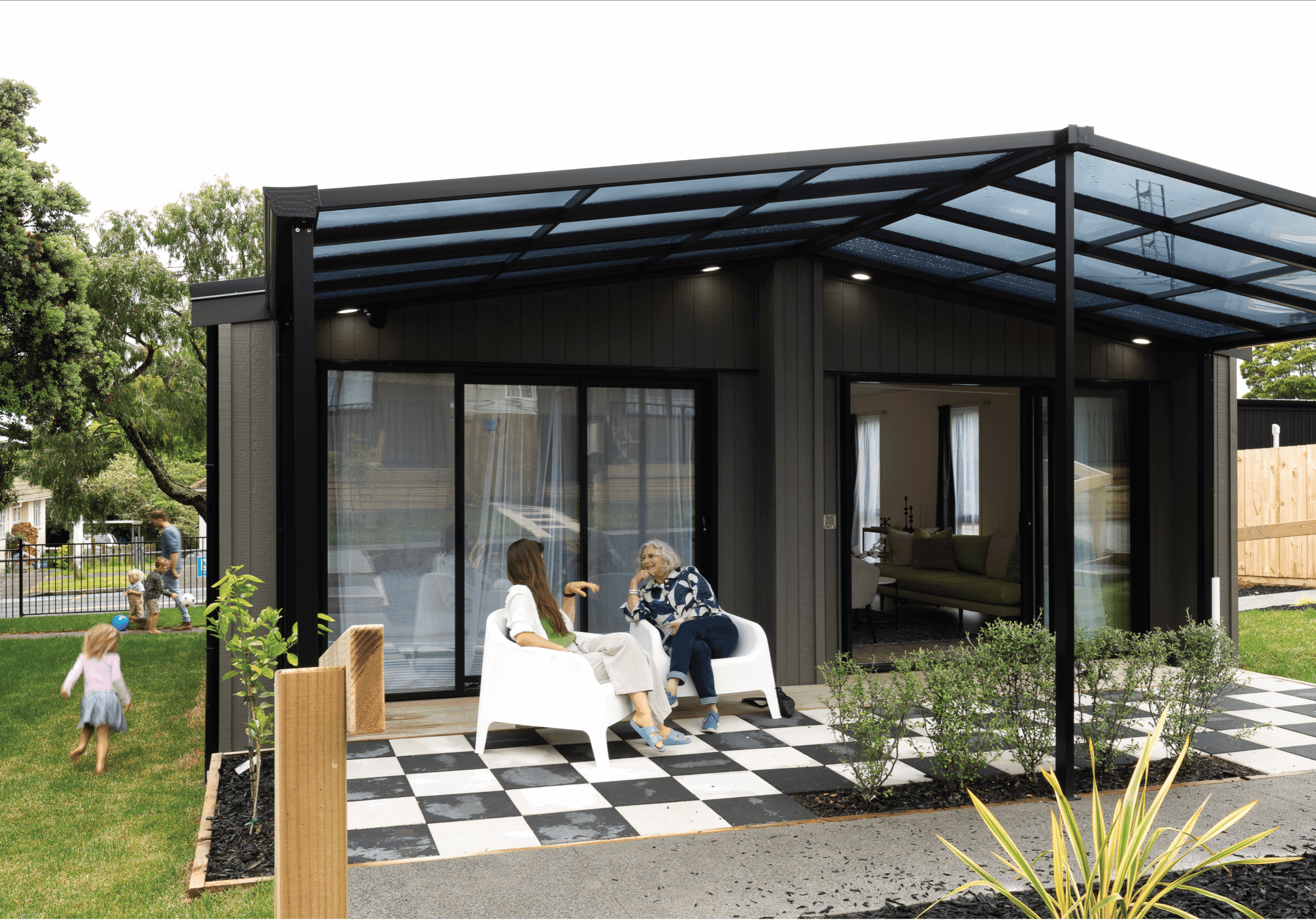 Photo with a mum and grandma speaking under a pergola outside an ecopod home, while a child runs towards her dad outside.  